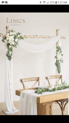 a wooden table topped with white flowers and greenery next to a chair covered in an arch