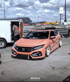 an orange car is parked in front of a trailer with the door open and two men working on it