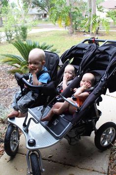 three babies are sitting in a stroller on the side walk, one is looking at the camera