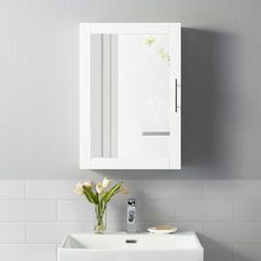 a white sink sitting under a bathroom mirror next to a wall mounted cabinet and vase with flowers