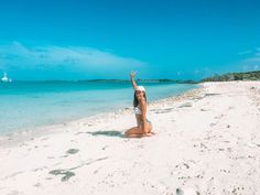 a woman sitting on top of a sandy beach next to the ocean with a sailboat in the background