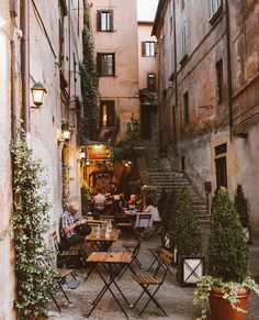 people sitting at tables in an alleyway with potted plants on the side walk