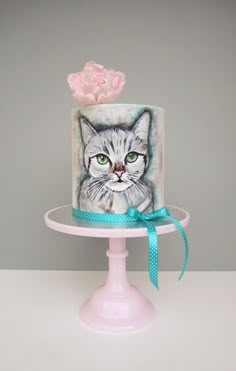 a cake decorated with a cat and a pink flower on top of a stand in front of a gray wall