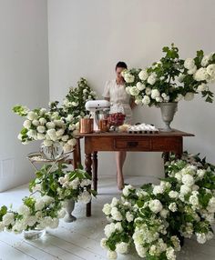 a woman sitting at a table surrounded by white flowers and potted plants on either side