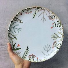 a hand holding a white plate with green leaves and berries painted on the bottom, in front of a gray background