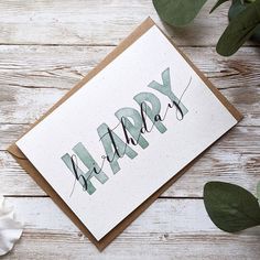 a card with the word happy on it next to some flowers and greenery in front of a white wooden background