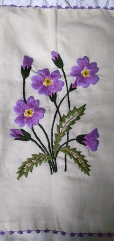 some purple flowers are on a white cloth with green stems and yellow centers, in the middle of an embroidered design