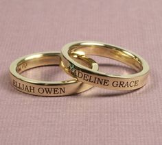 two gold wedding rings with names engraved on the sides, sitting on a pink background