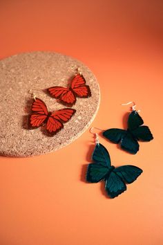 two red and black butterfly earrings on an orange surface next to a round earring