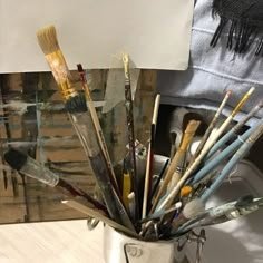 a cup filled with lots of paintbrushes on top of a white table next to a painting easel