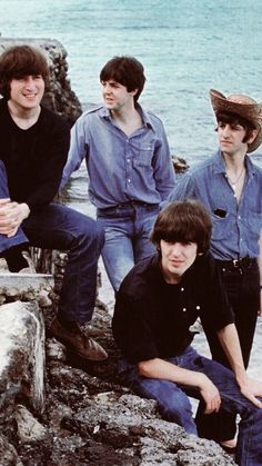 four young men are sitting on rocks near the water and one is wearing a hat