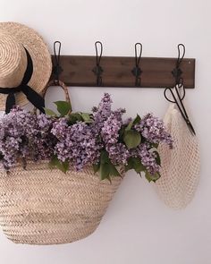 purple flowers in a basket hanging from a coat hook on a white wall next to a straw hat