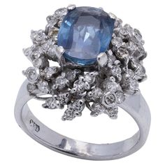 18kt white gold blue Ceylon sapphire diamond cluster ring. Design and Pattern: Design: The ring is centred with a large sapphire, surrounded by a cluster of round-cut diamonds, arranged in a floral or petal-like formation. Period: 1960s to 1970s Finger Size: UK L 1/2 (EU 51.5, US 6.25) Weight: 7.51 grams Diamonds - Cut: Round brilliant Quantity: 60 Carat weight: 1.20 carats in total Clarity: SI1 Colour: F - G Sapphire - Cut: Oval faceted Quantity:1 Carat weight: Approx. 2.00 carats in total Treatment: No treatment indicated. Condition: The ring is pre - owned, minor signs of usage, excellent condition overall. Luxury Sapphire Cubic Zirconia Cluster Ring, Luxury Sapphire Diamond Cluster Ring, Vvs Clarity Sapphire Cluster Ring, Luxury Blue Multi-stone Cluster Ring, Luxury Multi-stone Sapphire Cluster Ring, Ceylon Sapphire, Diamond Cluster Ring, Diamond Cluster, 1 Carat