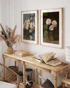 two framed pictures hang on the wall above a wooden table with books and flowers in vases