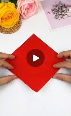 two hands holding up a red piece of paper next to flowers and pictures on a table