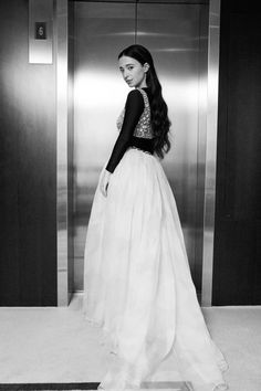 a woman standing in front of an elevator wearing a long white dress and black gloves