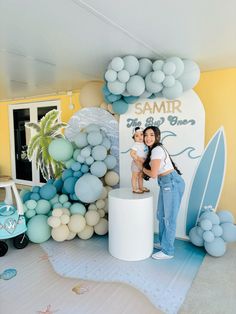 a woman holding a baby standing next to a surfboard on top of a table