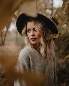 a woman with blonde hair wearing a black hat and grey sweater standing in the woods