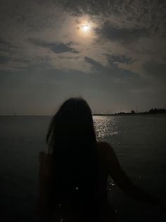 a woman standing in the water at night with her back turned to the camera and full moon behind her