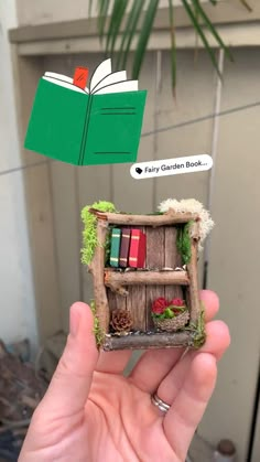 a hand holding up a miniature book shelf with books in it and an open book above it