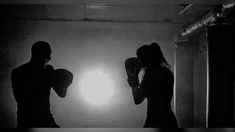 two people standing in a dark room with boxing gloves on their hands and one person holding a punching bag