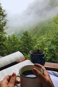 a person holding a cup of coffee and reading a book on a deck with mountains in the background