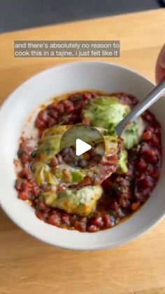 a person holding a spoon in a bowl filled with beans and broccoli on top of a wooden table