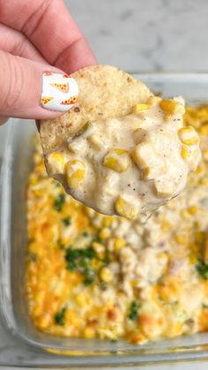 a hand holding a cracker over a casserole dish with corn and cheese