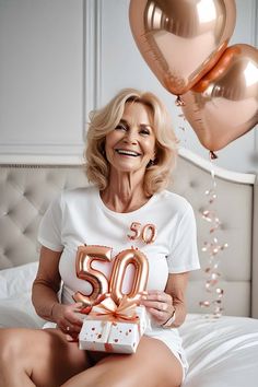 an older woman sitting on a bed holding a present box and balloons in the air