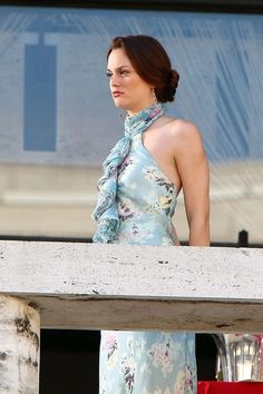 a woman in a floral dress standing on a balcony looking off into the distance with her hands behind her back