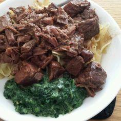 a white plate topped with pasta and beef next to spinach on top of a wooden table