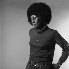 a woman with an afro standing in front of a white wall wearing jeans and a turtle neck sweater