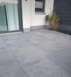 an empty concrete patio in front of a house with plants on the side and doors open