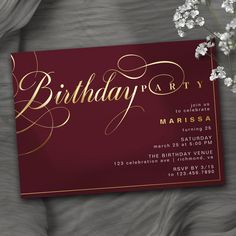 a red birthday party card with gold foil lettering and white flowers on the table next to it