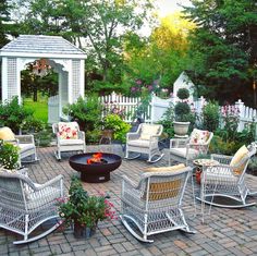 a patio with white wicker chairs and fire pit surrounded by greenery, shrubs and flowers