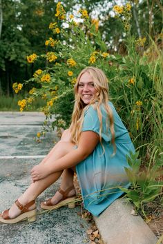 a woman sitting on the side of a road next to tall grass and yellow flowers