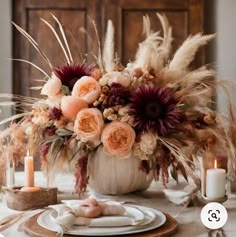 an arrangement of flowers and feathers on a table with candles in front of the centerpiece