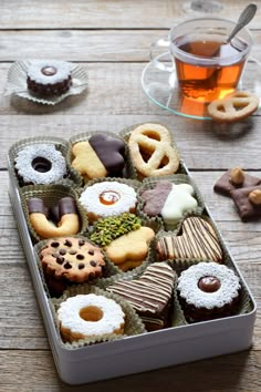 a tin filled with assorted pastries next to a cup of tea on top of a wooden table