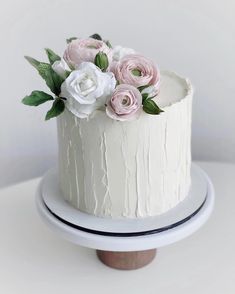 a white cake with pink and white flowers on top is sitting on a stand in front of a white wall