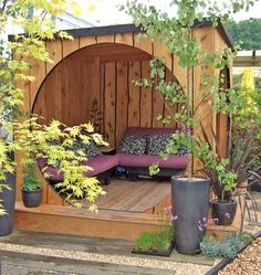 a small wooden shelter with purple cushions and pillows on the outside, surrounded by potted plants