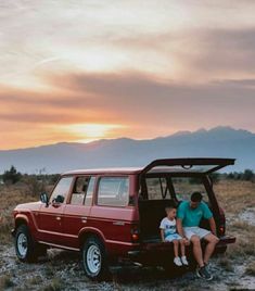 a man and child sitting in the back of a red truck on top of a dry grass field