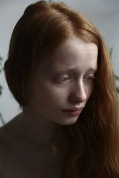 a woman with red hair and white makeup looks at the camera while standing in front of a plant