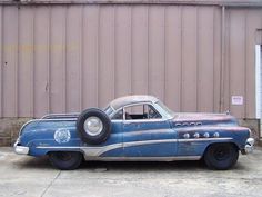 an old blue car parked in front of a building with a tire on it's side