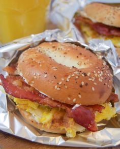 two sandwiches with bacon, eggs and cheese are on aluminum foil next to a glass of orange juice