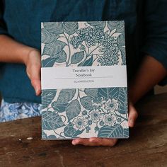 a person holding a book in their hands on top of a wooden table with blue and white flowers