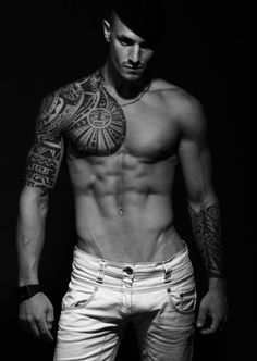 a shirtless man with tattoos on his chest and arms, standing in white jeans