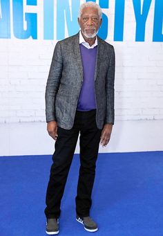 an older man standing in front of a white brick wall wearing a gray jacket and black pants