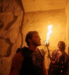 two men dressed in roman costumes stand next to each other, holding torches and looking into the distance