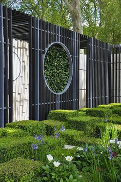 a garden with hedges and flowers in the foreground, surrounded by a circular metal fence