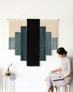 a woman sitting on a stool in front of a wall hanging with black and white fringes